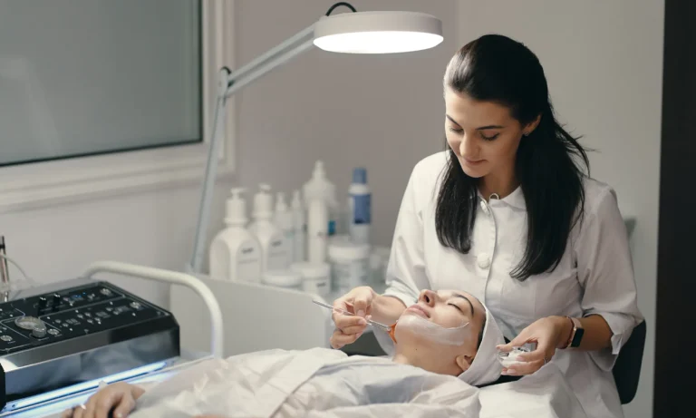 Female dermatologist performing a facial acne treatment on a patient lying on a clinic bed with skincare products and equipment nearby.