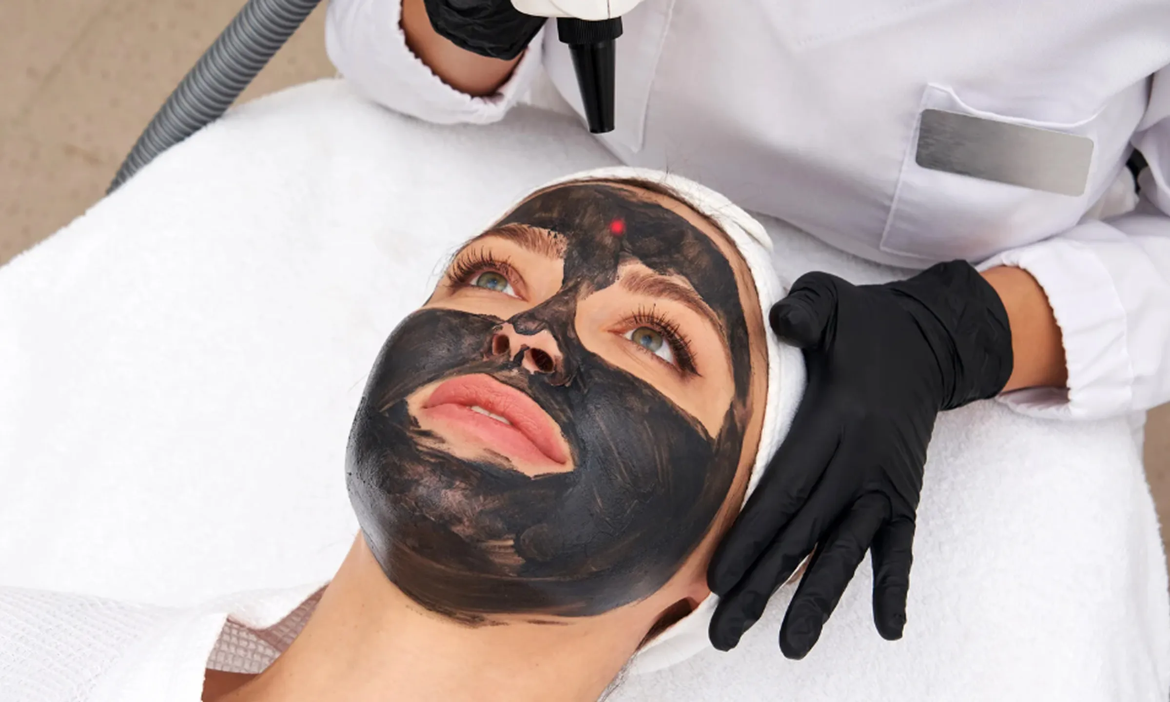 A woman undergoing a carbon facial treatment with a black carbon mask applied to her face while a dermatologist uses a laser device.